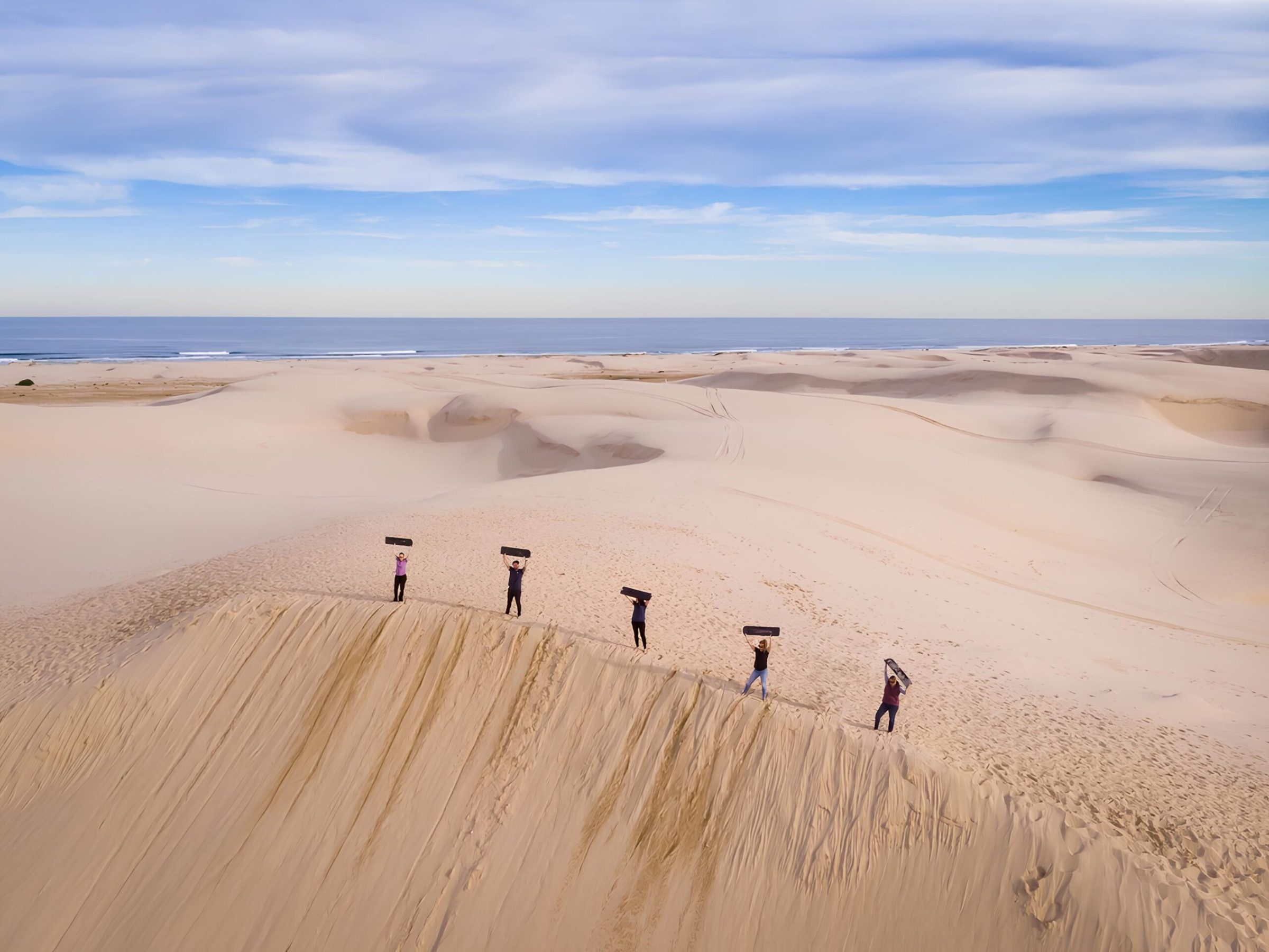 a group of people posing for the camera with their sandboards raised above their heads in anna bay, nsw